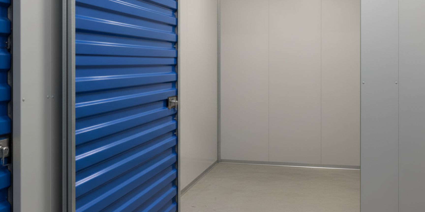 Großaufnahme eines einzelnen, abschließbaren Selfstorage-Abteils mit einer blauen Rolltür. Die Tür ist leicht geöffnet und gibt den Blick auf den leeren, sauberen Lagerraum frei, der bereit für die Einlagerung persönlicher Gegenstände oder kleinerer Geschäftswaren ist. Die Wand der Lagerbox ist in einem neutralen Grauton gehalten, was die Zweckmäßigkeit und den Schutz des Inhalts betont.